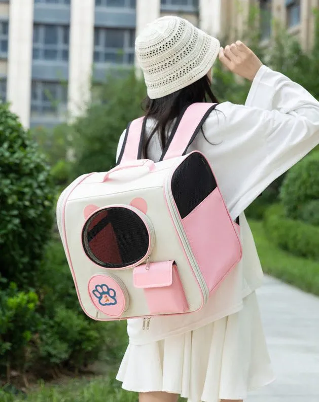 A woman wearing a white knitted hat and white clothing carries a pink and beige pet backpack with cat ear - shaped circular mesh window and paw print detail, standing on a pathway amidst greenery and buildings outdoors.