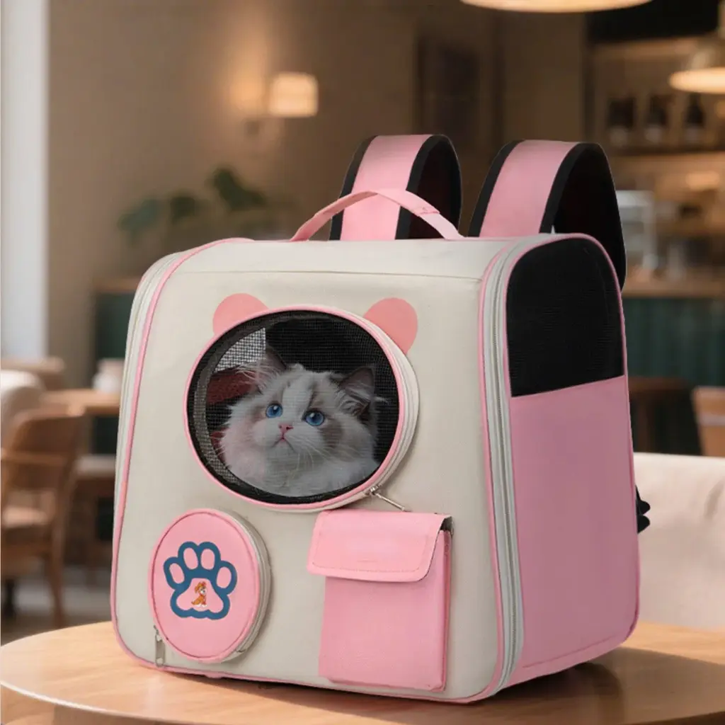 A cute pink and beige pet carrier backpack with cat ear details and a circular mesh window, through which a Ragdoll cat with striking blue eyes is visible. The backpack has a paw print - themed pocket and is placed on a wooden table in a cozy indoor space, possibly a café.